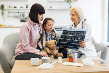 Bir doktor bir anne ve çocuğuna röntgen filmi gösterir, battaniyeye sarılmış bir şekilde, tıbbi danışma ortamında..