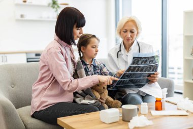 Bir doktor, tıbbi muayene sırasında anne ve çocuğuna röntgen çeker ve sonuçları ve tedavi seçeneklerini tartışır..