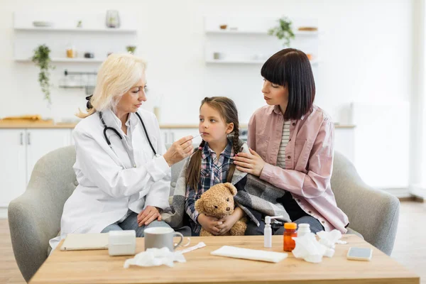 Bir doktor termometreyle genç bir kızı muayene ederken annesi onu rahatlatır, evde bakım ve ilgi gösterir..