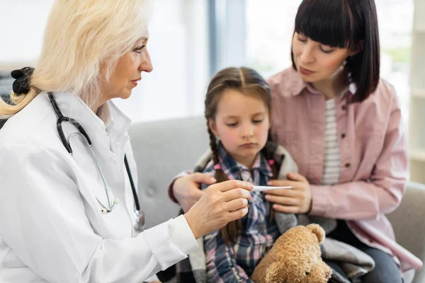 Bir doktor, annesi yanında olan genç bir kızı hastalığını teşhis etmek için termometreyle muayene eder..
