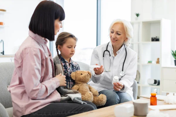 Bir doktor battaniyeye sarılmış ve muhtemelen bir klinikte oyuncak ayı tutan bir anneye ve kızına ilaç gösteriyor..