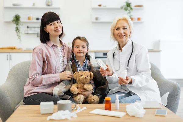 Bir doktor evde bir anneye ve hasta kızına danışmanlık yapıyor, çocuğa ilaç ve bakım öneriyor. Kız battaniyeye sarılmış..