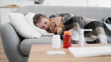Genç bir kız kanepede uyuyor, oyuncak ayıyla sarılıp yatıyor, yanında ilaçla, hastalık ve istirahat öneriyor..