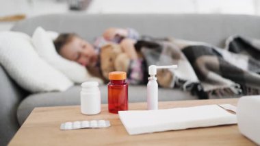 Küçük bir çocuk kanepede yatıyor, hasta gibi görünüyor, öndeki masada ilaç ve mendillerle..