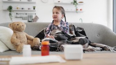 Battaniyeye sarılı genç bir kız hasta ve evinde bir kanepede otururken boğazını tutuyor gibi görünüyor..