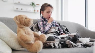 Genç bir kız, battaniyeye sarılmış, bir kanepede oyuncak ayıyla otururken öksürüyor..
