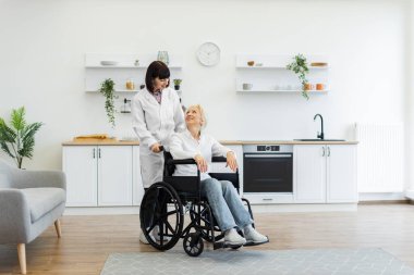 Gülümseyen bir bakıcı, yaşlı bir kadını tekerlekli sandalyedeki parlak, modern bir mutfaktan geçirir. Rahatlık ve bakım duygusu yaratır..