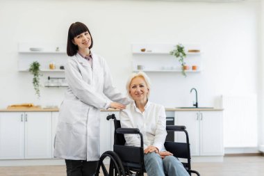 Beyaz önlüklü genç bir kadın tekerlekli sandalyedeki yaşlı bir kadının yanında duruyor, ikisi de gülümsüyor. Parlak ve temiz bir mutfak ortamındalar..