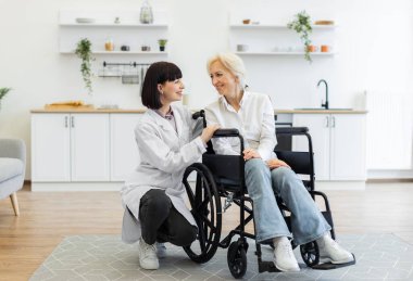 Laboratuvar önlüğü giymiş genç bir bakıcı, parlak, modern bir evde tekerlekli sandalyedeki yaşlı bir kadına yardım ederken gülümsüyor..