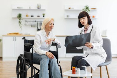Bir doktor tekerlekli sandalyedeki yaşlı bir hastaya röntgeni açıklıyor. Parlak bir odadalar, küçük bir masada ilaç tutuyorlar..