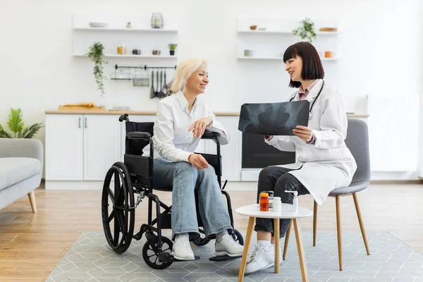 Bir doktor, tekerlekli sandalyede oturan gülümseyen yaşlı bir kadına röntgen filmi gösteriyor. Tıbbi muayenede sonuçları tartışıyorlar..