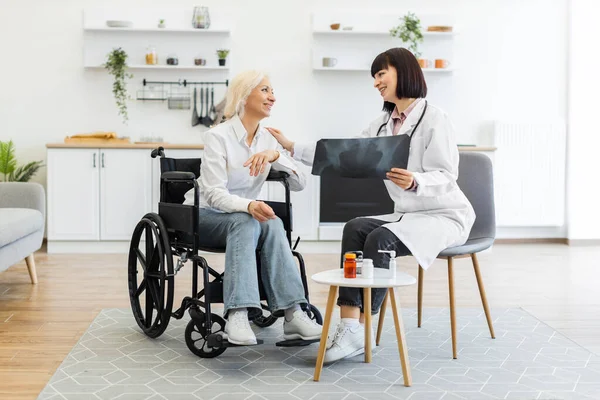 Kadın bir doktor, tekerlekli sandalyede oturan yaşlı bir kadına röntgen filmi gösteriyor. Parlak bir odada sonuçları tartışıyor..
