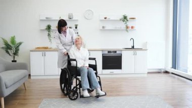 Bir bakıcı, modern bir mutfakta, tekerlekli sandalyedeki yaşlı bir kadına yardım eder. Bakım ve destek sahnesi yaratır..