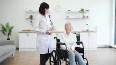 Beyaz önlüklü bir doktor, parlak ve modern bir odada tekerlekli sandalyede oturan yaşlı bir kadınla konuşuyor..