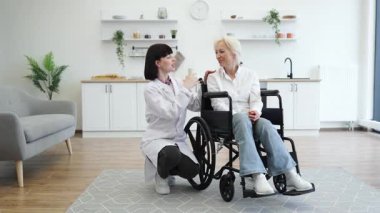 Genç bir bakıcı, her ikisi de parlak bir mutfak ortamında sıcak bir şekilde gülümseyen tekerlekli sandalyedeki yaşlı bir kadına destek olur. Sahne bakım ve desteği iletir..