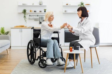 Kadın bir doktor, tekerlekli sandalyedeki yaşlı bir kadına ilaç veriyor. Ev ortamında tedavi hakkında konuşuyor..