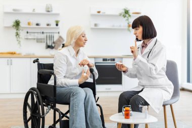 Kadın bir doktor, tekerlekli sandalyedeki yaşlı bir hastaya, elinde haplar ve masada bir şişeyle ilaç vermeyi açıklar..