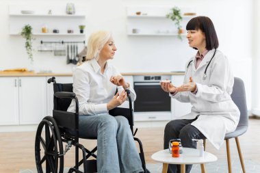 Beyaz önlüklü kadın bir doktor, tekerlekli sandalyedeki yaşlı bir kadına ev ortamında tedavi seçeneklerini tartışan ilaçları açıklıyor..