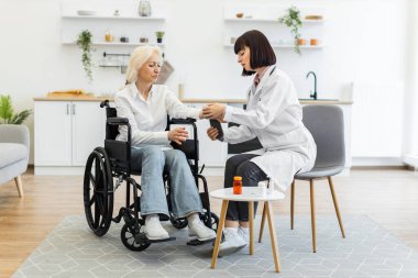 Kadın bir doktor, tekerlekli sandalyedeki yaşlı bir kadının bileğini muayene eder. Muhtemelen ev ortamında sağlığını ya da yaralanmasını değerlendirir..