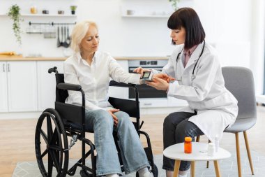 Beyaz önlüklü bir kadın doktor, tekerlekli sandalyede oturan yaşlı bir kadının kan basıncını ölçer..