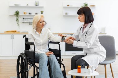 Kadın bir doktor, tıbbi muayene odasında tekerlekli sandalyede oturan yaşlı bir kadının kan basıncını ölçer. Hasta sıradan kıyafetler giyiyor..