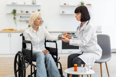 Kadın bir doktor, ev ziyareti sırasında tekerlekli sandalyedeki yaşlı bir kadının kan basıncını ölçer..