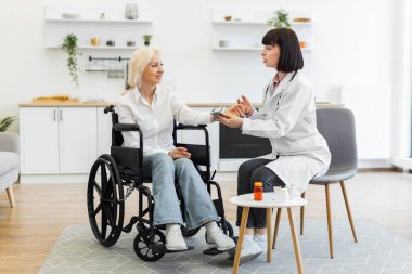 Kadın bir doktor, yaşlı bir kadının kan basıncını ölçer. Evde tekerlekli sandalyede oturmuş, sağlığını tartışırken..