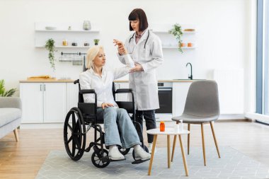 Beyaz önlüklü kadın bir doktor, yaşlı bir kadının kolunu incelerken o da parlak, modern bir mutfakta tekerlekli sandalyede oturuyor..