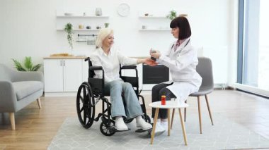 Beyaz önlüklü bir kadın doktor, tekerlekli sandalyede oturan yaşlı bir kadının kan basıncını ölçer..
