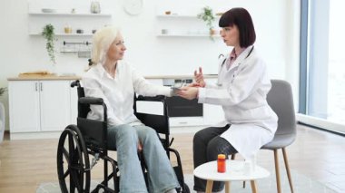 Kadın bir doktor, tekerlekli sandalyedeki yaşlı bir kadının kan basıncını ölçer. Parlak, modern bir ev ortamında sağlığını tartışır..