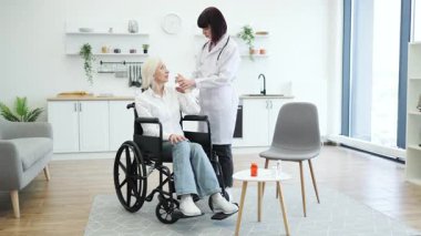Bir doktor yaşlı bir kadının kolunu muayene eder. Tekerlekli sandalyede otururken, muhtemelen bir ev ortamında..