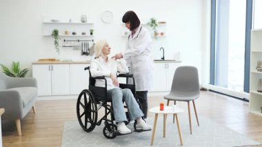Kadın bir doktor, tekerlekli sandalyedeki yaşlı bir kadına kol egzersizlerinde yardımcı olur. Ev ortamında rehabilitasyonu ve iyileşmeyi teşvik eder..