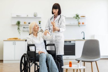 Beyaz önlüklü bir kadın doktor tekerlekli sandalyede oturan yaşlı bir kadının kolunu muayene ediyor, muhtemelen hareket ya da ağrı var mı diye kontrol ediyor..