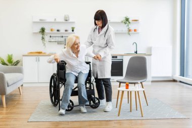 Doktor, yaşlı bir kadına tekerlekli sandalyeden kurtulması için yardım eder..