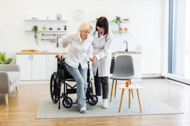 Kadın bir doktor, yaşlı bir kadının hareket kabiliyetini ve bakımını arttırmak için iyi ışıklandırılmış bir odada tekerlekli sandalyesinden kalkmasına yardım eder..