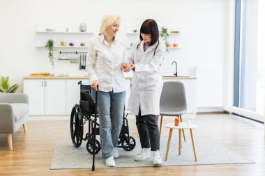 Kadın bir doktor, yaşlı bir kadının tekerlekli sandalyesiyle yürümesine yardım ediyor. Bir evin içindeler..