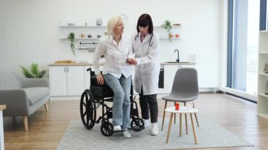 Kadın bir doktor, yaşlı bir kadının tekerlekli sandalyeden kalkmasına yardım eder. Bağımsızlığı ve bakımı teşvik eder. Sahne iyi aydınlatılmış, modern bir evde geçiyor..