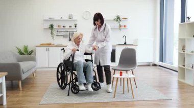 Kadın bir doktor, yaşlı bir kadına yardım eder. Parlak bir odada, tekerlekli sandalyeden kalkar ve ona destek olur..