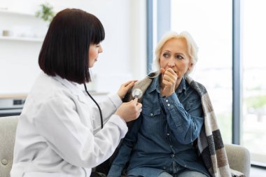 Kadın bir doktor, yaşlı bir kadını muayene etmek için steteskop kullanıyor. Öksürüyormuş gibi görünüyor..