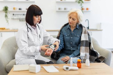 Kadın bir doktor, yaşlı bir kadının kan basıncını ölçerken bir kanepede, battaniyeye sarılmış olarak oturuyor. Tıbbi malzemeler masada..