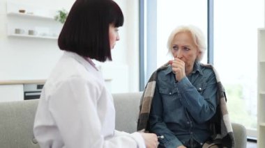 Kadın bir doktor, yaşlı bir kadını muayene etmek için steteskop kullanıyor. Öksürüyormuş gibi görünüyor..