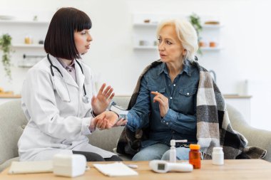 Beyaz önlüklü kadın bir doktor, koltukta otururken yaşlı bir kadını muayene ediyor ve sağlığını tartışıyor. Hasta battaniyeye sarılı..