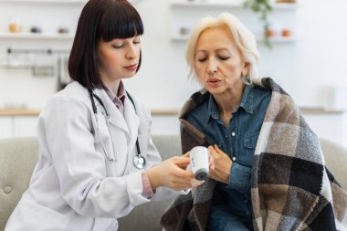 Bir doktor dijital termometre kullanan yaşlı bir kadının sıcaklığını kontrol ediyor. Kadın battaniyeye sarılmış..