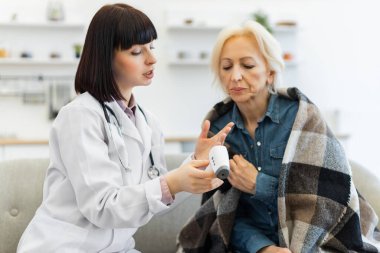 Kadın bir doktor yaşlı bir kadına hasta battaniyeye sarılı iken dijital termometreyi nasıl kullanacağını gösteriyor..