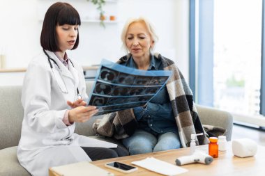 Beyaz önlüklü kadın bir doktor yaşlı bir kadına röntgen filmini açıklar. Muhtemelen teşhis ve tedavi seçeneklerini evde tartışırlar..
