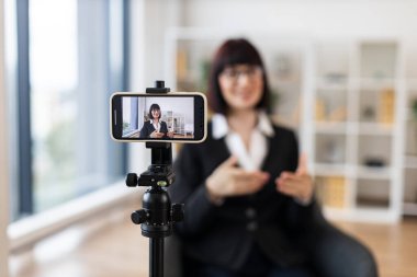 Beyaz kadın resmi giyinmiş, tripoda monte edilmiş akıllı telefon kullanarak profesyonel bir iş blogunu kaydediyor, el hareketleriyle kavramları ifade ediyor, iletişimi teşvik ediyor.