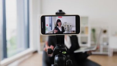 Profesyonel giyimli beyaz kadın dizüstü bilgisayarı ve kamerasıyla blog yazıyor, iş kavramlarını tartışıyor. Ofis arka planı profesyonel atmosfer taşır.