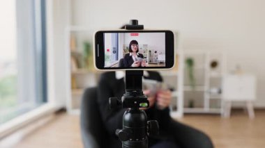 30 'lu yaşların ortalarında beyaz profesyonel bir kadın yatırımlar ve nakit paralar hakkında video blogu kaydediyor, tripoda monte edilmiş akıllı telefon kamerasıyla modern çalışma ortamını gösteriyor.