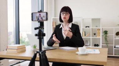 Beyaz kadın gözlüklü ve siyah ceketli. Masada kameralı iş blogu var.