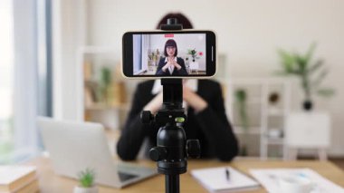 Takım elbiseli beyaz bir kadın ofis masasında oturmuş akıllı telefonlu profesyonel video kaydediyor. Sahne, iletişim, işyeri modernliği, çevrimiçi ilişki ve bilgi kavramlarını öneriyor.